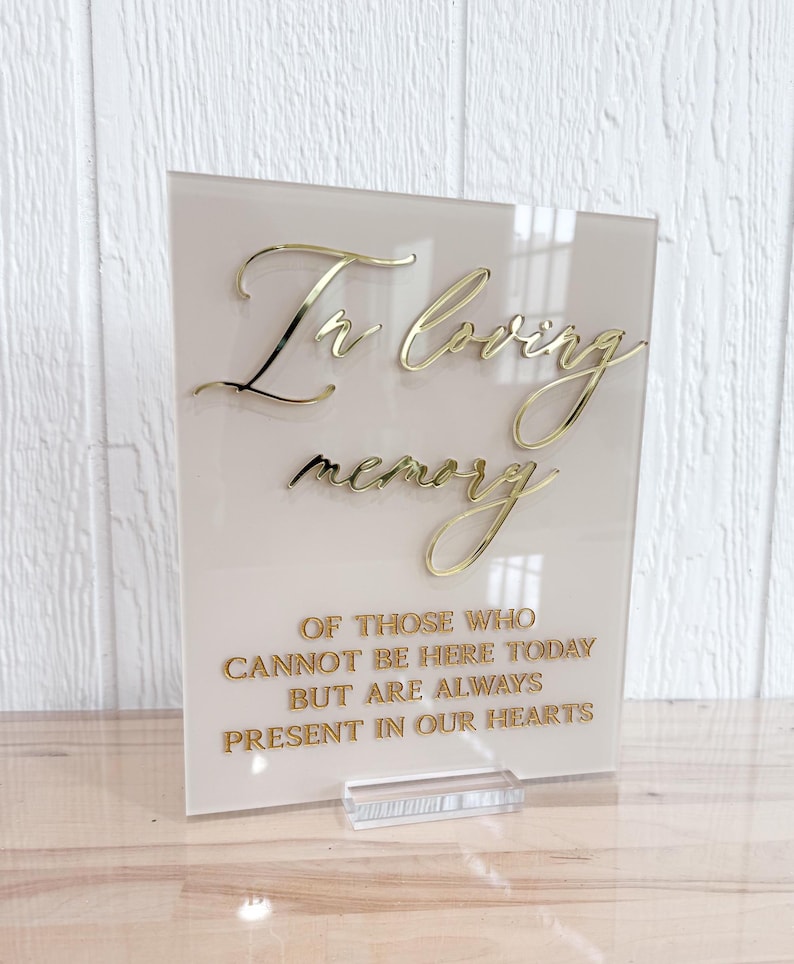 May include: A white acrylic sign with gold lettering that reads "In loving memory of those who cannot be here today but are always present in our hearts".