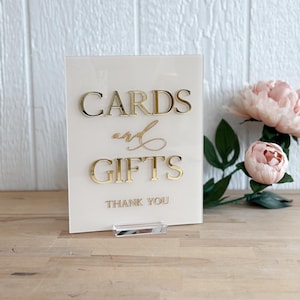 May include: A white acrylic sign with gold lettering that reads "Cards and Gifts" with the words "Thank You" below. The sign is on a wooden surface.