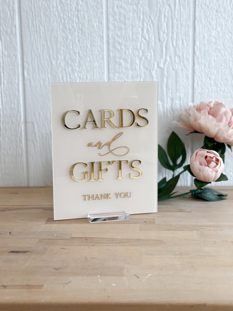 May include: A white acrylic sign with gold lettering that reads "Cards and Gifts" with the words "Thank You" below. The sign is on a wooden surface.