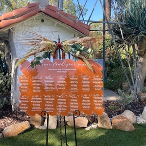 May include: A clear acrylic wedding seating chart with a brown, orange, and white abstract design. The chart lists table numbers and guest names. The text reads "Please find your seat", "Kourtney and Austin Santara", and "09/16/2022".