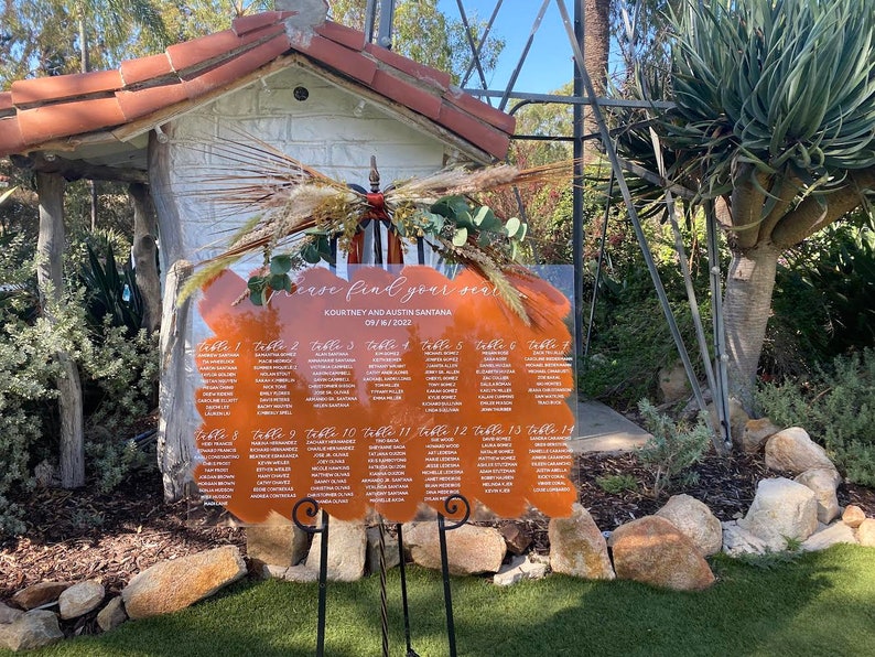 May include: A clear acrylic wedding seating chart with a brown, orange, and white abstract design. The chart lists table numbers and guest names. The text reads "Please find your seat", "Kourtney and Austin Santara", and "09/16/2022".
