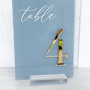 May include: A blue-grey table number sign with the word "table" in white script. A gold number "4" is affixed to the sign. The sign is supported by a clear acrylic base. Ideal for weddings and events.