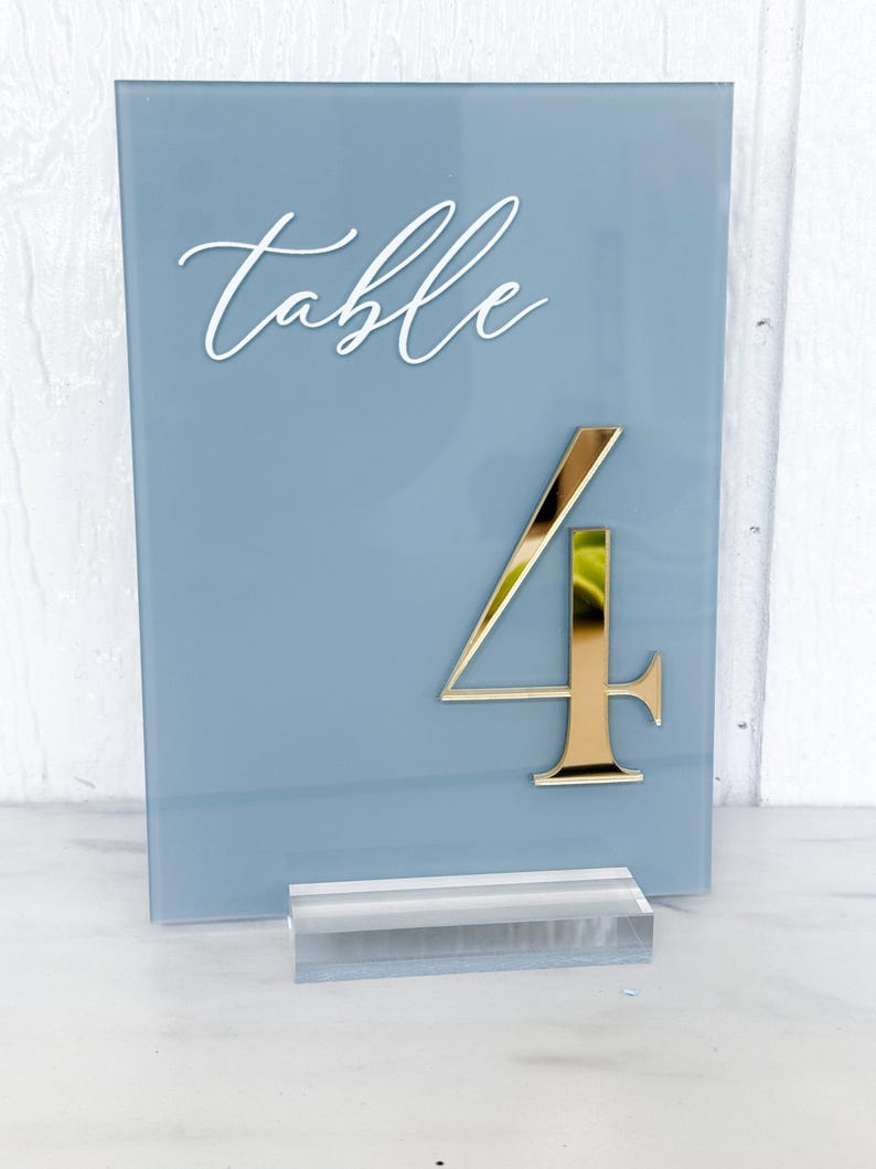 May include: A blue-grey table number sign with the word "table" in white script. A gold number "4" is affixed to the sign. The sign is supported by a clear acrylic base. Ideal for weddings and events.