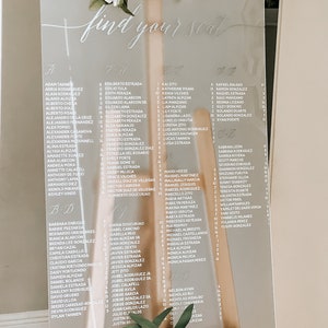 May include: A clear acrylic seating chart with white lettering. The chart is titled "Find Your Seat" and lists names in alphabetical order. The chart is on a wooden easel.