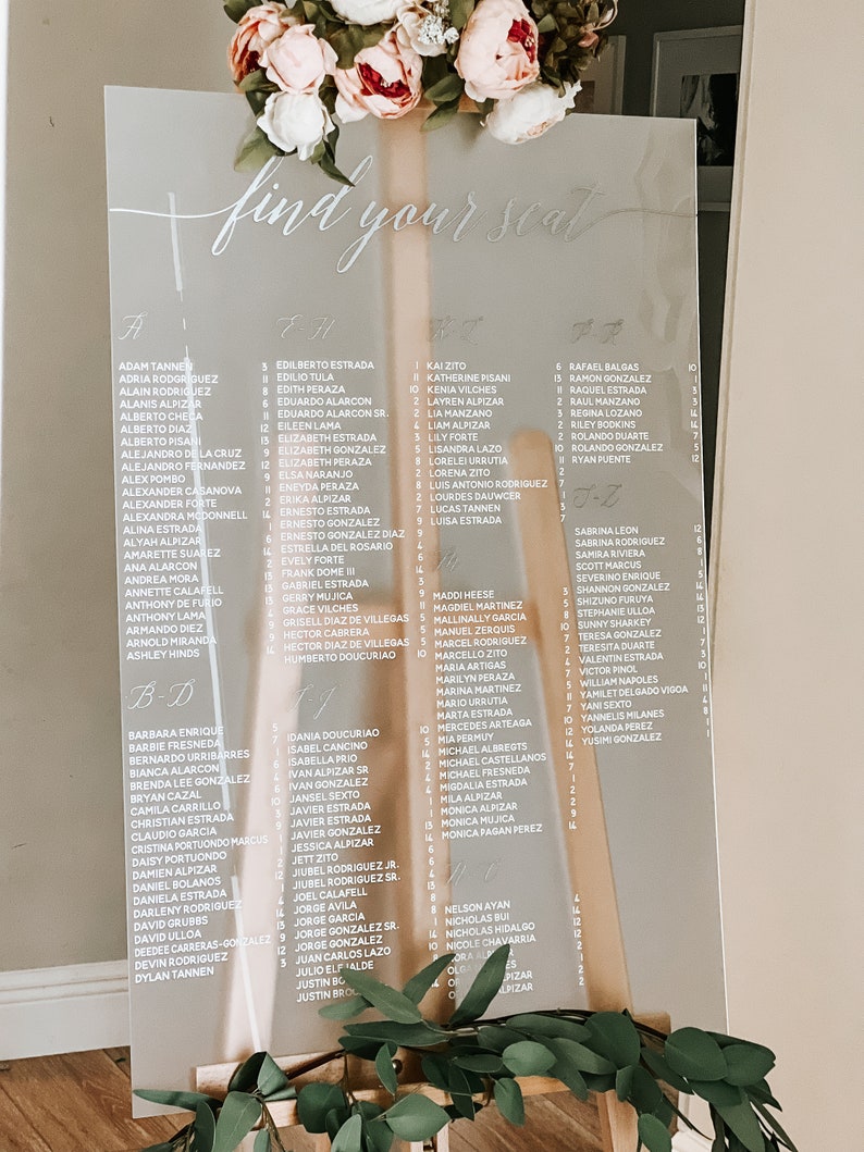 May include: A clear acrylic seating chart with white lettering. The chart is titled "Find Your Seat" and lists names in alphabetical order. The chart is on a wooden easel.