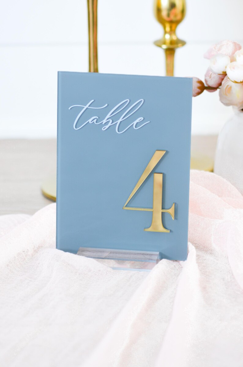 May include: A blue table number with the word "table" in white script and a gold number 4. The table number is displayed on a white and pink cloth. The table number is for a wedding or event.