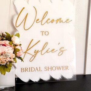 May include: White acrylic sign with gold lettering that reads "Welcome to Kylie's Bridal Shower". The sign has a white brushstroke design.