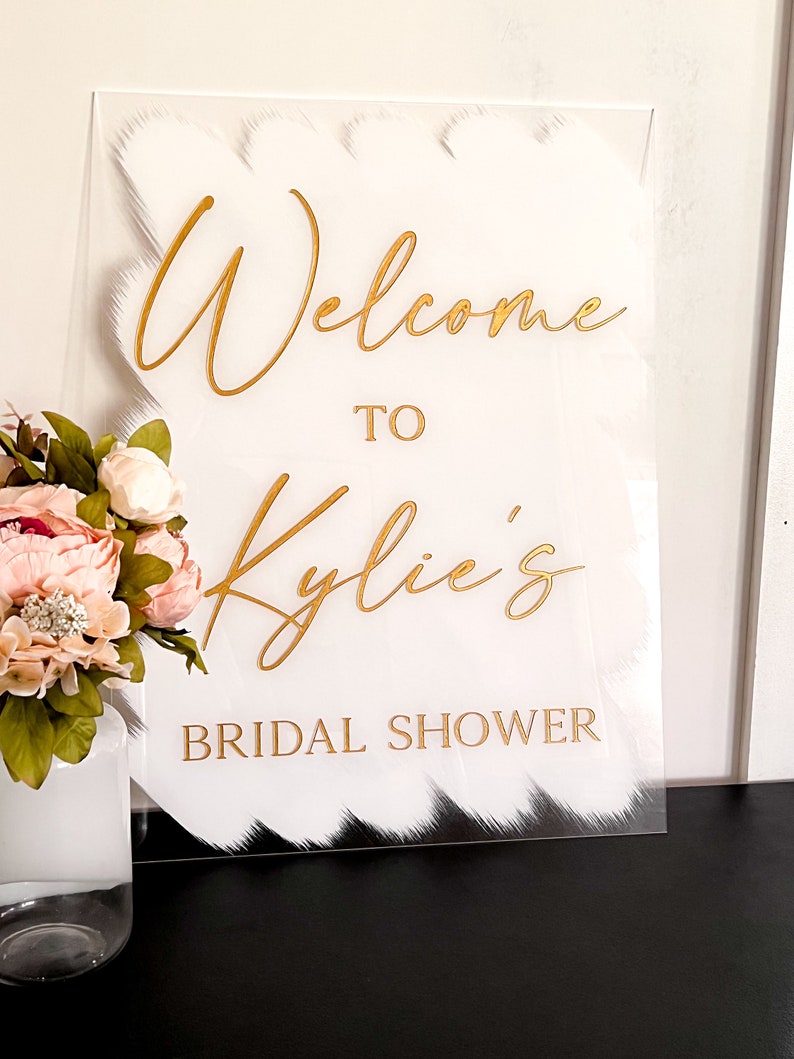 May include: White acrylic sign with gold lettering that reads "Welcome to Kylie's Bridal Shower". The sign has a white brushstroke design.