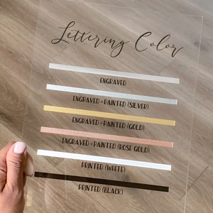 May include: A clear acrylic sign with the text "Lettering Color" at the top. The sign lists different lettering options with corresponding colour swatches: Engraved, Engraved + Painted (Silver), Engraved + Painted (Gold), Engraved + Painted (Rose Gold), Printed (White), and Printed (Black).