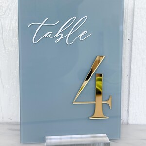 May include: A blue-grey table number with the word "table" in white script. A gold number "4" is affixed to the front. The sign is displayed on a clear acrylic base.