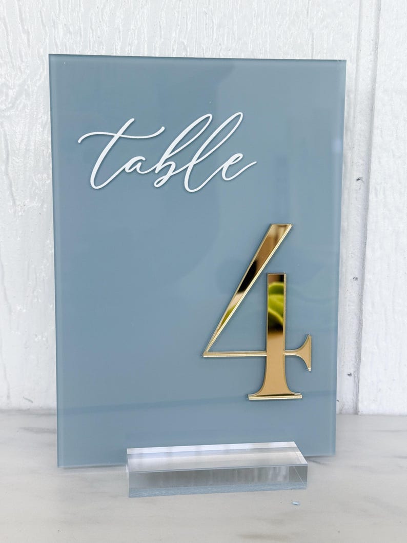 May include: A blue-grey table number with the word "table" in white script. A gold number "4" is affixed to the front. The sign is displayed on a clear acrylic base.