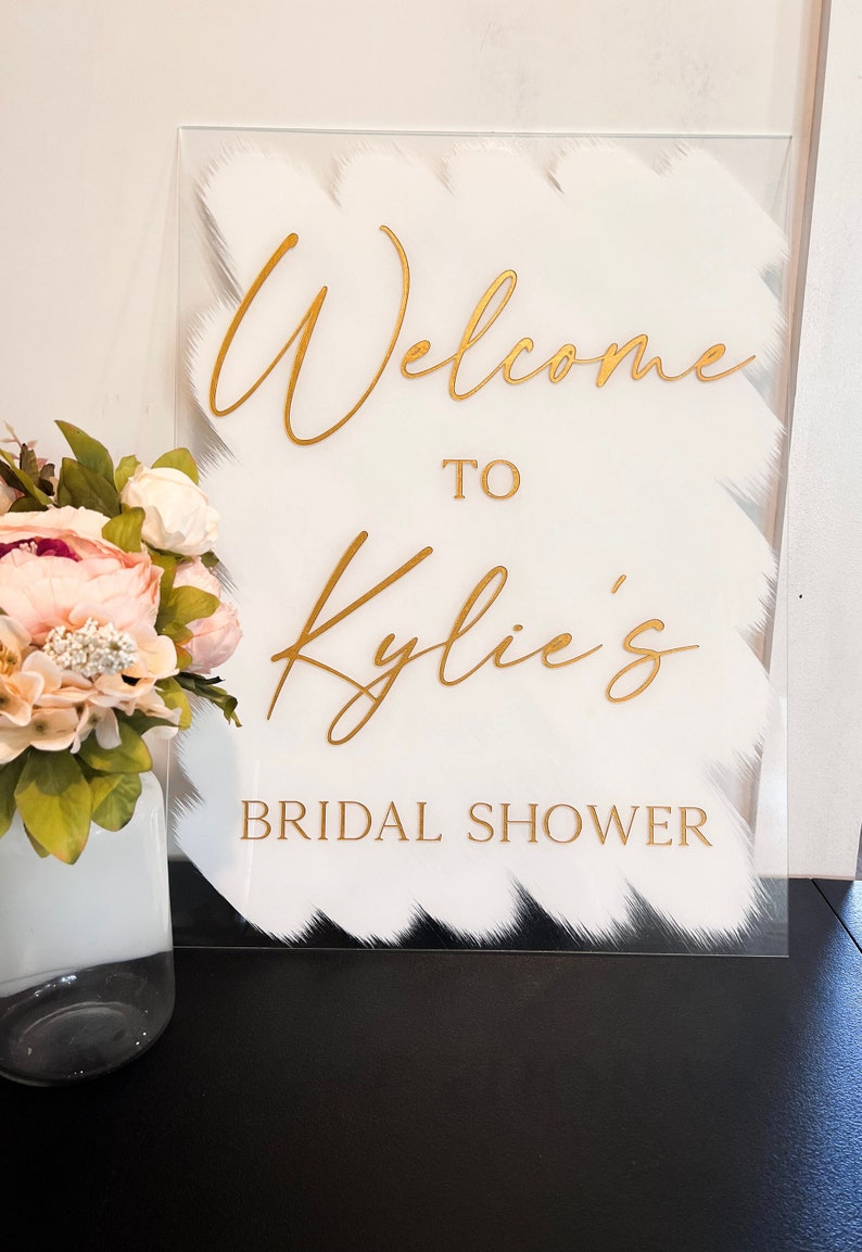 May include: White acrylic sign with gold lettering that reads "Welcome to Kylie's Bridal Shower".
