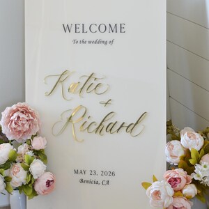 May include: A cream-colored wedding welcome sign with gold lettering that reads "Welcome" and "Katie & Richard." The date "May 23, 2026" and location are also displayed. Floral arrangements frame the sign.
