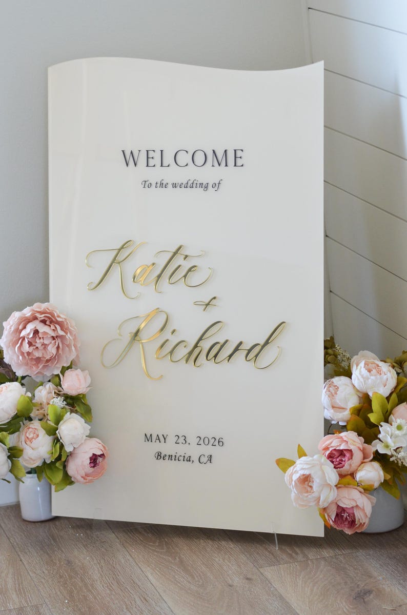 May include: A cream-colored wedding welcome sign with gold lettering that reads "Welcome" and "Katie & Richard." The date "May 23, 2026" and location are also displayed. Floral arrangements frame the sign.