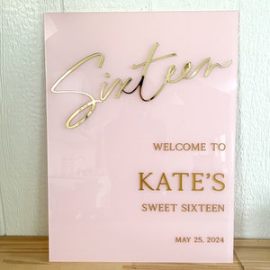 May include: Pink acrylic sign with gold lettering that says "Sixteen" in cursive, "Welcome to Kate's Sweet Sixteen" and "May 25, 2024".