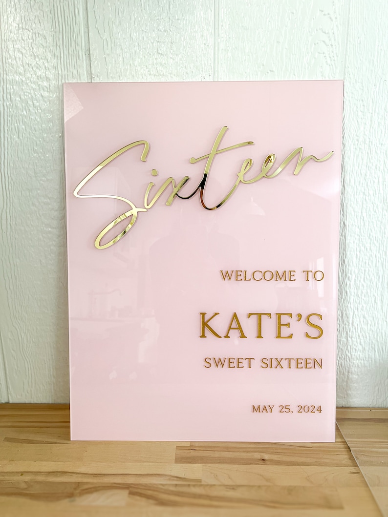 May include: Pink acrylic sign with gold lettering that says "Sixteen" in cursive, "Welcome to Kate's Sweet Sixteen" and "May 25, 2024".