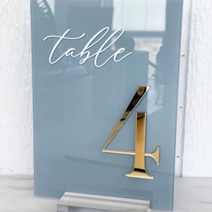 May include: A blue-grey acrylic table number with the word "table" in white script at the top. A gold number "4" is affixed to the front. The sign is displayed with a clear acrylic base.