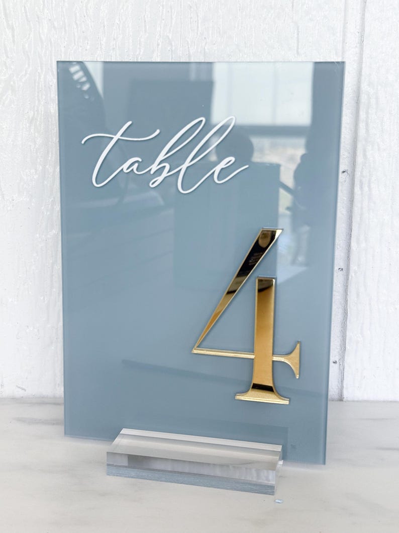 May include: A blue-grey acrylic table number with the word "table" in white script at the top. A gold number "4" is affixed to the front. The sign is displayed with a clear acrylic base.