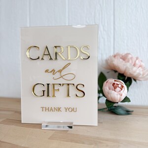 May include: A white acrylic sign with gold lettering that reads "Cards and Gifts" with the words "Thank You" below. The sign is on a clear acrylic stand and is sitting on a wooden surface.