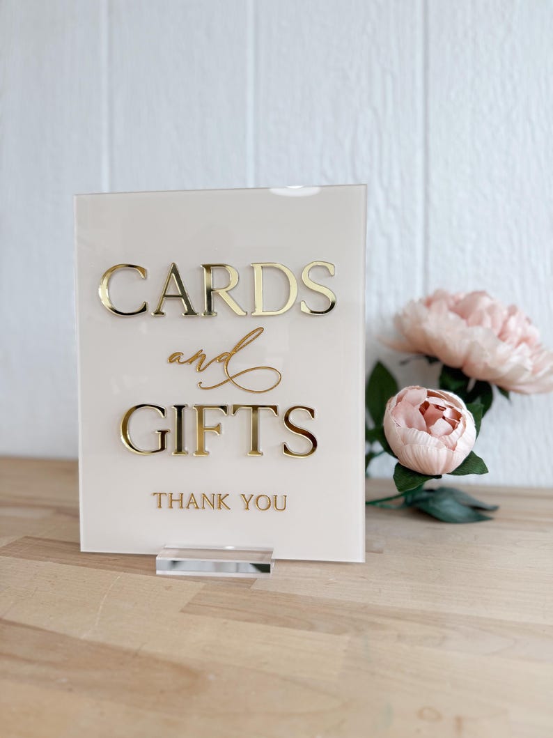 May include: A white acrylic sign with gold lettering that reads "Cards and Gifts" with the words "Thank You" below. The sign is on a clear acrylic stand and is sitting on a wooden surface.