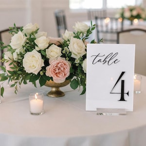 May include: A table setting with a floral arrangement and candles. A white sign with the word "table" and the number "4" is on the table. The flowers are white and pink roses in a gold vase.