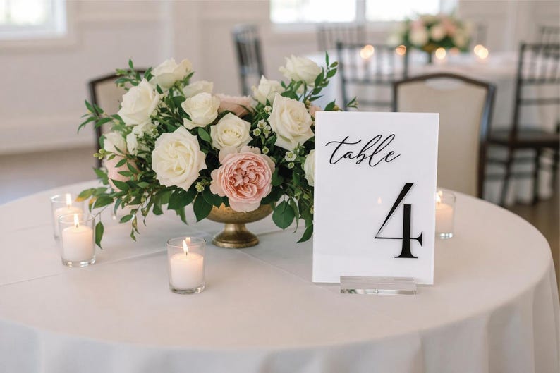 May include: A table setting with a floral arrangement and candles. A white sign with the word "table" and the number "4" is on the table. The flowers are white and pink roses in a gold vase.