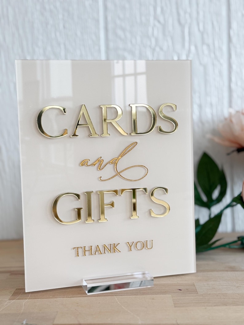 May include: A white acrylic sign with gold lettering that reads "Cards and Gifts Thank You".
