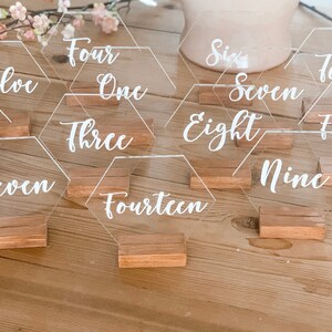 May include: A set of 15 clear acrylic hexagon table numbers with white calligraphy. Each number is on a small wooden stand. The numbers range from one to fifteen.