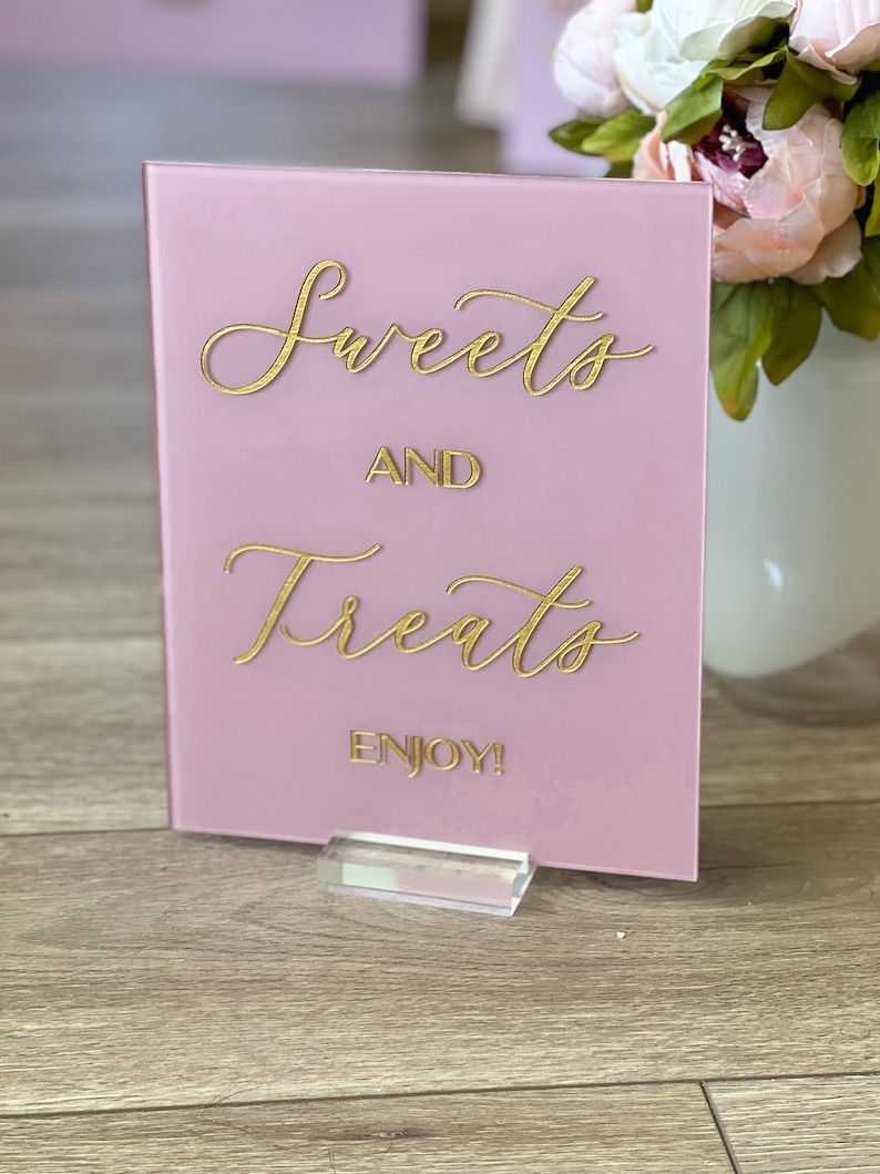 May include: A pink acrylic sign with gold lettering that reads "Sweets and Treats" and "Enjoy!". The sign is held up by a clear acrylic stand. A vase of flowers is in the background.