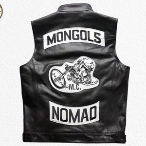 Könnte beinhalten: Schwarze Lederweste mit weißen Aufnähern. Der obere Aufnäher lautet "MONGOLS", der untere "NOMAD", und ein zentraler Aufnäher zeigt einen Biker auf einem Motorrad. Die Weste hat einen klassischen Biker-Stil.