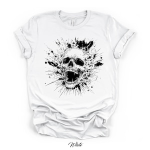 May include: A white short-sleeve t-shirt featuring a central black and white graphic print of a human skull with an open jaw, surrounded by dynamic ink splatter and splash effects that radiate outward from the center of the chest area.