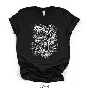 May include: A black short-sleeve t-shirt featuring a white graphic print of a stylized, abstract skull design. The artwork consists of sharp, jagged lines and splattered ink effects that form the facial structure of a skull on the chest area.