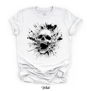 May include: White t-shirt featuring a black and white graphic of a screaming skull with an explosion-like design. The word "White" is written in cursive at the bottom of the shirt. The shirt has a crew neck and short sleeves.