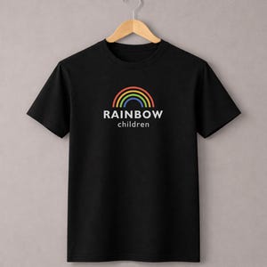Könnte beinhalten: Schwarzes T-Shirt mit einem Regenbogen-Design und dem weißen Schriftzug "RAINBOW children". Der Regenbogen hat rote, orange, gelbe, grüne und blaue Bögen. Das T-Shirt hängt an einem Holzkleiderbügel.