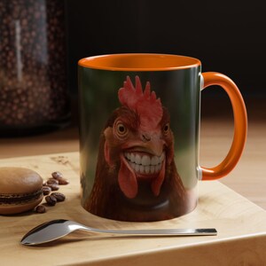 Könnte beinhalten: Eine Keramik-Kaffeetasse mit orangefarbenem Innenraum und Griff. Die Tasse zeigt eine Nahaufnahme des Gesichts eines Huhns mit einem menschenähnlichen Lächeln. Die Tasse steht auf einer Holzoberfläche mit einem Löffel und einem Gebäck.