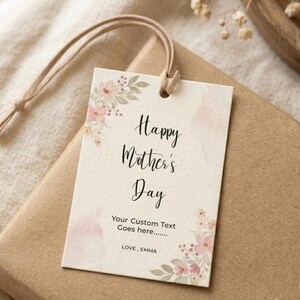 May include: A gift tag with the text "Happy Mother's Day" and space for custom text, adorned with watercolor floral accents. The tag is tied with a beige string and rests on a wrapped gift.