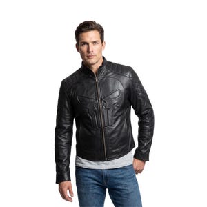 May include: A black leather jacket featuring a prominent embossed skull emblem on the chest, a full-length front zipper, a stand-up collar, and quilted detailing on the shoulders. The jacket is worn over a light gray t-shirt and blue jeans.
