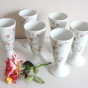Puede incluir: Un juego de cinco copas de porcelana blanca con un delicado diseño floral de pequeñas enredaderas rosas, amarillas y verdes. Una rosa fresca con pétalos rojos, rosas y amarillos se encuentra en primer plano, mostrando hojas verdes naturales.