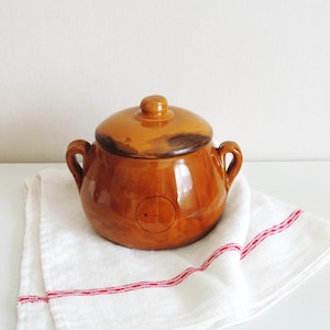Puede incluir: Una olla de cerámica marrón vidriada de forma redonda, con dos asas laterales y una tapa a juego. La olla presenta un acabado liso y brillante con una hendidura circular en la parte frontal, sobre un paño blanco con rayas rojas finas.