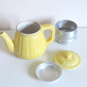 Op de afbeelding: Een gele keramische theepot met een geribbelde buitenkant, een bijpassend deksel met knop, een zilverkleurig metalen filterinzetstuk en een apart zilverkleurig metalen cilindrisch onderdeel. De theepot heeft een gebogen tuit en een stevig handvat.