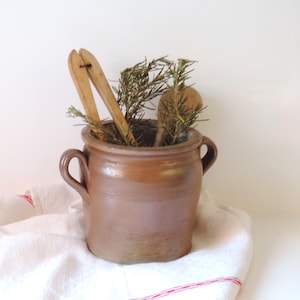 Może przedstawiać: Brązowy ceramiczny garnek z dwoma bocznymi uchwytami, zawierający drewniane szczypce kuchenne i drewnianą łyżkę oraz gałązki zielonego rozmarynu. Garnek ma gładkie, szkliwione wykończenie i stoi na białej ściereczce z czerwonym paskiem.