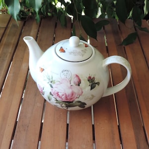 Peut inclure: Une théière en céramique blanche ornée d'un motif floral composé de grandes roses roses, de feuilles vertes et de petits papillons orange. Le corps de la théière présente un motif de tampon postal circulaire. Couvercle assorti avec bouton.