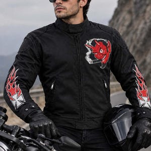Könnte beinhalten: Eine schwarze Motorradjacke mit einem roten Teufelsmotiv auf der linken Brust sowie roten Flammenmustern und weißen Kreuzen auf beiden Ärmeln. Die Jacke verfügt über einen Stehkragen, einen zentralen Reißverschluss und zwei Reißverschlusstaschen.