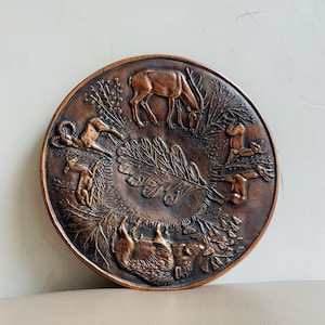 May include: A round, bronze-colored decorative plate with a relief design featuring various animals and foliage. The plate's rim and center are darker, with a lighter bronze hue highlighting the raised details of the animals and plants. The animals include deer, a ram, and other wildlife.