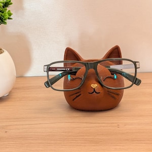 Peut inclure: Un porte-lunettes en forme de chat marron avec des oreilles pointues, un petit nez en forme de cœur doré et des moustaches peintes. Une paire de lunettes rectangulaires gris foncé à monture fine repose sur le devant du support en forme de chat.