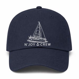 May include: Navy blue baseball cap featuring a white embroidered sailboat design. The cap includes the text "N' JOY CREW" in white, with an anchor symbol. The hat has a curved brim and an adjustable strap.