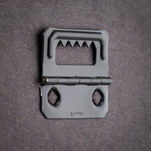 May include: Black metal picture hanging hook with a hinged design and a series of teeth on the top. The hook has two holes on the bottom and the text "18.71.20054" is visible.
