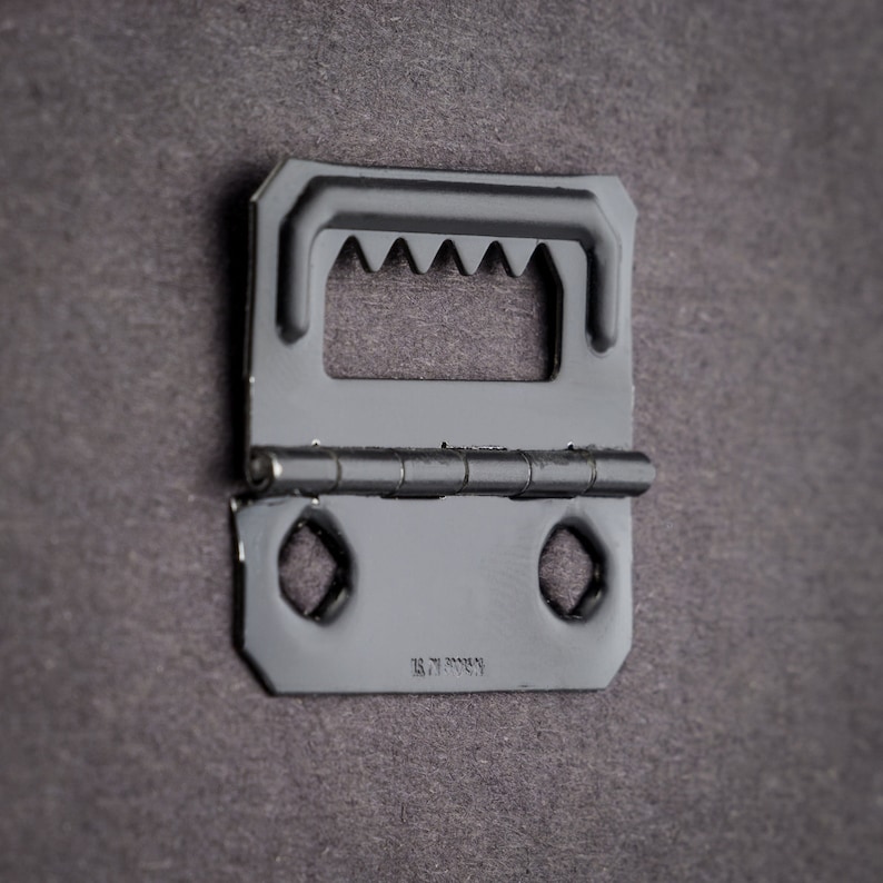 May include: Black metal picture hanging hook with a hinged design and a series of teeth on the top. The hook has two holes on the bottom and the text "18.71.20054" is visible.