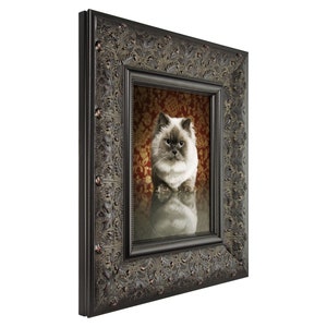 May include: A framed portrait of a fluffy cat with a serious expression. The cat has light-colored fur with darker points. The ornate, dark brown frame features detailed floral and scrollwork designs. The background is a patterned red and gold.