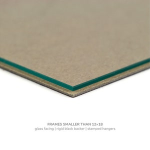 May include: Close-up of the edge of a picture frame showing a glass facing, a rigid black backer, and stamped hangers. FRAMES SMALLER THAN 12x18.